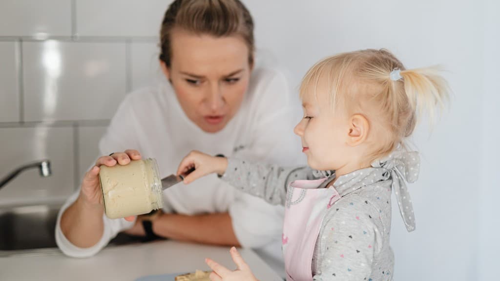 A parent watching closely as a toddler holds a jar and a knife in the kitchen